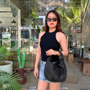 May include: A black sleeveless ribbed knit top, a light blue denim mini skirt, and a black textured hobo bag with a snakeskin pattern. The outfit is paired with dark-tinted cat-eye sunglasses featuring a black frame and gold-tone accents.