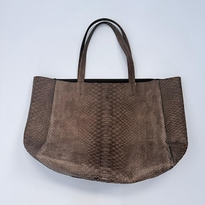 May include: A large tote bag in a muted brown tone featuring a textured snakeskin pattern. The bag has two thin, matching brown shoulder straps and a wide, rectangular body with a distinct central panel of larger, vertical scales.