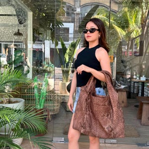 May include: A large, slouchy hobo bag featuring a brown snakeskin-textured pattern. The bag has a wide shoulder strap, a tassel detail hanging from the side, and a spacious, unstructured silhouette made from a flexible material with a glossy finish.