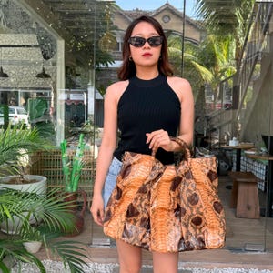 May include: A large tote bag featuring a textured snakeskin pattern in shades of tan, orange, and dark brown. The bag has two sturdy top handles and a spacious, slouchy silhouette with a visible scale texture throughout the exterior surface.