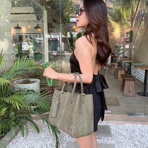 May include: A sage green tote bag with a textured snakeskin pattern, featuring two top handles and a matching tassel charm. The bag has a structured rectangular shape and is crafted with a detailed scale-like surface texture throughout.