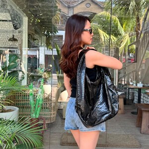 Puede incluir: Un bolso hobo negro con un patrón texturizado de piel de serpiente, que presenta una silueta holgada y una correa ancha para el hombro. El bolso está fabricado con un material brillante con un diseño de escamas repetitivo en negro.