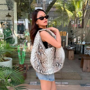 Könnte beinhalten: Eine große Schultertasche im Hobo-Stil mit strukturiertem Schlangenledermuster in Beige-, Braun- und Weißtönen. Die Tasche hat eine lockere, unstrukturierte Silhouette mit einem breiten, integrierten Riemen und einem detaillierten Schuppenmuster.