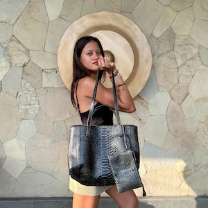 May include: A black tote bag with a textured snakeskin pattern, featuring two long shoulder straps and a matching smaller rectangular zippered pouch attached to the front. The bag has a structured rectangular shape with a glossy finish.