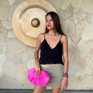 Puede incluir: Una blusa negra tipo camisola con tirantes finos y escote en V combinada con una minifalda verde oliva lisa y un bolso de mano tipo sobre texturizado en color rosa intenso. El conjunto se complementa con varias pulseras en ambas muñecas.