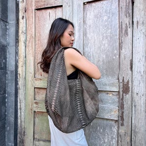 Könnte beinhalten: Eine große, lässige Hobo-Tasche mit strukturiertem, dunkelbraunem Schlangenledermuster. Die Tasche verfügt über einen breiten, integrierten Schulterriemen und eine weiche, unstrukturierte Silhouette, die natürlich fällt. Das Material zeigt ein detailliertes Schuppenmuster.
