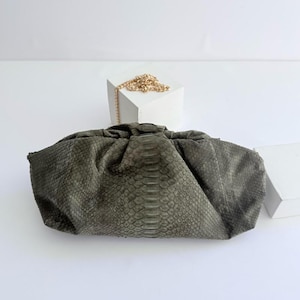 Op de afbeelding: Een donker olijfgroene clutch met een textuur van slangenleer. De tas heeft een geplooid, zacht ontwerp met een centraal verticaal paneel en een dunne goudkleurige metalen kettingriem die op een wit blok achter de tas rust.