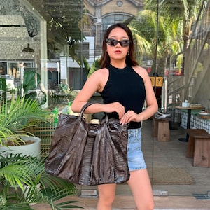 May include: A large, dark brown snakeskin-textured tote bag with a slouchy, pleated silhouette, two top handles, and a central vertical panel. The bag features a glossy finish and a structured opening, showcasing a detailed reptilian scale pattern.