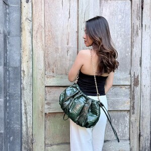 May include: A dark green, textured snakeskin-patterned shoulder bag with a gathered, rounded silhouette. The bag features a metallic gold-tone chain strap and a matching snakeskin-patterned wrist strap, providing a versatile carrying option.