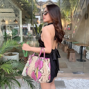 May include: A black satin halter-style top with a tie-back closure paired with a structured handbag featuring a vibrant pink, yellow, and natural snakeskin-patterned leather exterior, dual top handles, and a decorative tassel accent.