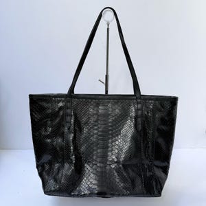 Op de afbeelding: Een zwarte tote bag met een gestructureerd slangenleerpatroon aan de buitenkant. De tas heeft twee dunne zwarte schouderbanden aan de bovenrand en een gestructureerde rechthoekige basis met verticale stiksels op het voorpaneel.