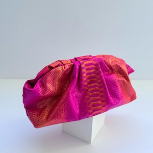 May include: A vibrant fuchsia and orange clutch bag featuring a textured snakeskin pattern. The bag has a gathered, soft-structured design with a central vertical panel and a horizontal top closure, showcasing a bright gradient colour effect.