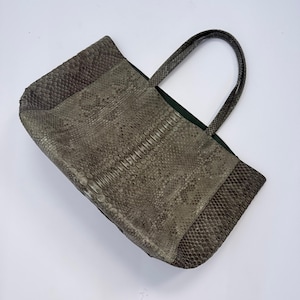 Puede incluir: Un bolso tote rectangular con un patrón de piel de serpiente texturizado en tonos grises en el exterior. El bolso incluye dos asas a juego con el mismo patrón de piel de serpiente en la parte superior y un forro interior de color verde oscuro.