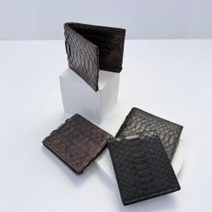 May include: A collection of three bi-fold wallets featuring a textured snakeskin pattern. One wallet is displayed open in a dark brown tone, while two closed wallets are shown in dark brown and black, showcasing the detailed scale-like surface.