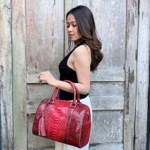 May include: A red snakeskin-patterned handbag with a cylindrical shape, two top handles, and a matching red tassel charm attached to one side. The bag features a textured scale-like surface and metallic hardware connecting the handles to the body.