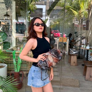 May include: A black sleeveless ribbed knit top, a light blue denim mini skirt, and a brown and black snakeskin patterned clutch bag. The clutch features a metallic zipper closure and a thin matching strap, held in front of the body.