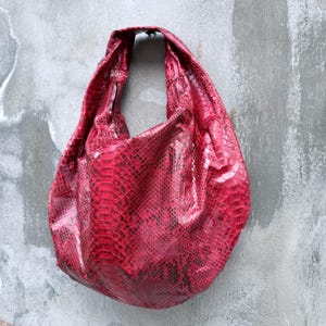 May include: A hobo-style shoulder bag featuring a vibrant red and black snakeskin-patterned texture. The bag has a slouchy, unstructured silhouette with a single integrated handle at the top, showcasing a detailed scale-like surface throughout.
