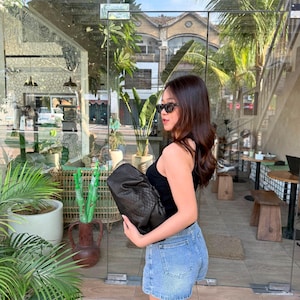 Puede incluir: Un bolso de mano tipo sobre de cuero negro texturizado con una silueta fruncida, llevado bajo el brazo. El bolso presenta una forma suave y voluminosa con un patrón tejido o en relieve visible en su superficie y un cierre de marco integrado.