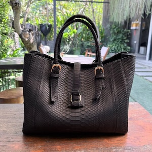 May include: A dark charcoal gray handbag with a textured snakeskin pattern. The bag features two top handles, a central vertical strap with a buckle closure, and two additional side straps with metallic buckles on the front panel.