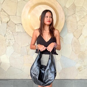 May include: A black hobo bag with a textured snakeskin pattern, featuring a slouchy, crescent-shaped silhouette and a wide shoulder strap. The bag is constructed from a glossy material with a detailed scale-like surface texture throughout.