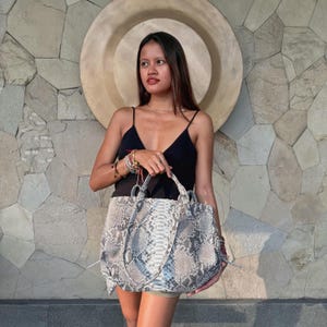 Può includere: Una borsa tote grande caratterizzata da un motivo a pelle di serpente grigio e bianco con finitura testurizzata. La borsa presenta due manici superiori e una forma rettangolare strutturata con soffietti laterali. Il materiale mostra squame.