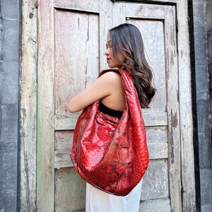 May include: A large, slouchy hobo-style shoulder bag featuring a vibrant red snakeskin-patterned texture. The bag has a wide, integrated strap that rests comfortably over the shoulder, showcasing a glossy finish and a distinct reptilian scale design.