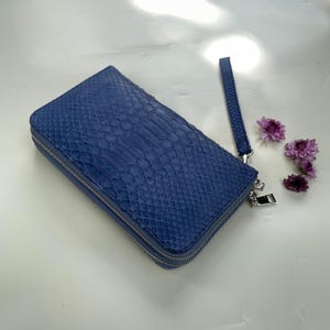 Puede incluir: Una cartera rectangular azul con un patrón texturizado de piel de serpiente, que cuenta con un cierre de cremallera de metal plateado y una correa de muñeca desmontable a juego. La cartera tiene un diseño estructurado con esquinas redondeadas.