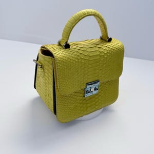 May include: A bright yellow handbag featuring a textured snakeskin pattern. The bag has a single top handle, a structured rectangular body, and a silver-toned metal clasp closure on the front flap. The sides of the bag are dark in color.