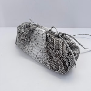 May include: A cylindrical pouch bag featuring a textured snakeskin pattern in shades of gray, white, and black. The bag has a gathered top closure secured with a thin, matching snakeskin-textured drawstring cord that ties into a small bow.