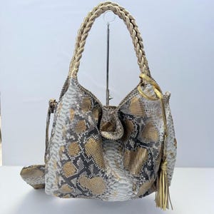 May include: A hobo-style handbag featuring a snakeskin pattern in shades of gold, white, and black. The bag includes a single braided gold handle and a gold tassel accent hanging from the side. The exterior displays a textured, scale-like finish.