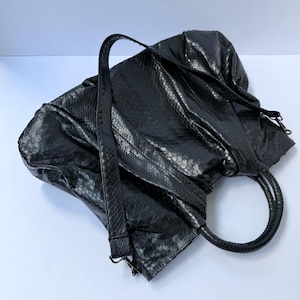 Könnte beinhalten: Eine schwarze Handtasche mit einem strukturierten Schlangenledermuster auf der gesamten Außenseite. Die Tasche verfügt über einen langen, verstellbaren Schulterriemen und zwei abgerundete Tragegriffe mit Metallbeschlägen an den Riemen.