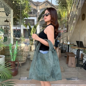May include: A large, dark green hobo-style shoulder bag with a textured snakeskin pattern, featuring a wide strap, a central vertical seam detail, and a decorative tassel hanging from the side attachment point of the bag strap.