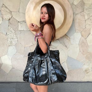 Puede incluir: Un bolso de mano grande y holgado con un acabado metálico de color gris oscuro con textura de piel de serpiente. El bolso tiene dos asas superiores resistentes, un diseño fruncido en la parte superior y una silueta espaciosa y sin estructura.