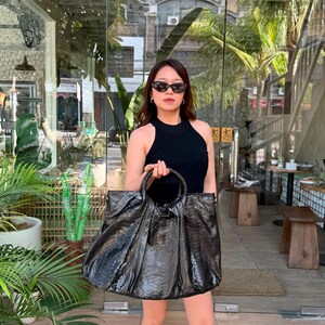 May include: A large, dark grey snakeskin-textured handbag with two rounded top handles and a gathered, slouchy silhouette. The bag features a metallic sheen and a spacious, unstructured design, showcasing a detailed reptilian scale pattern throughout.