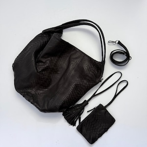 May include: A black hobo bag with a snakeskin texture, featuring two top handles, a detachable shoulder strap, and a matching small zippered pouch with a long tassel. The set is crafted with a consistent dark, reptilian-patterned finish throughout.