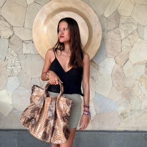 May include: A large tote bag featuring a brown and tan snakeskin pattern with a textured scale finish, two rounded top handles, and a spacious slouchy silhouette. The bag is constructed with a central vertical panel and side gussets.