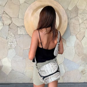 May include: A crossbody bag featuring a textured snakeskin pattern in shades of gray, white, and black. The bag has a wide, adjustable matching snakeskin strap, a curved top flap, and a black zippered front pocket with metallic hardware accents.