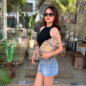 May include: A sleeveless black top paired with a light blue denim mini skirt and a textured snakeskin-patterned handbag with a gold chain strap. The handbag features a gradient of brown, tan, and cream scales in a structured, rectangular shape.
