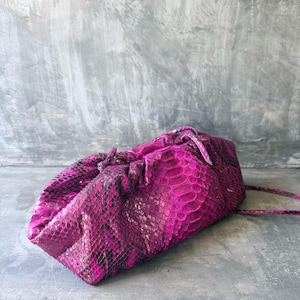 Puede incluir: Un bolso de mano con estampado de piel de serpiente en magenta y negro con un diseño de bolsa fruncida y suave. El bolso presenta una superficie texturizada con escamas y una correa fina a juego en el lateral, con un color vivo y audaz.
