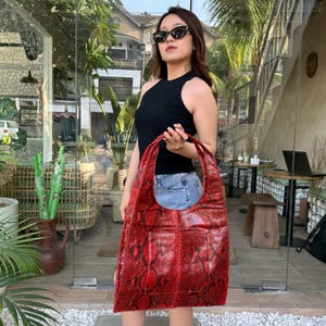 May include: A large hobo-style shoulder bag featuring a vibrant red and black snakeskin-patterned texture. The bag has a wide, integrated shoulder strap and a rectangular front pocket, constructed from a flexible material with a glossy finish.