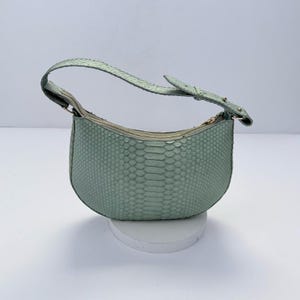 Könnte beinhalten: Eine halbmondförmige Handtasche in blassem Mintgrün mit einer strukturierten Schlangenhautoptik. Die Tasche verfügt über einen einzelnen verstellbaren Schulterriemen mit Schnalle und einen Reißverschluss an der Oberseite. Die Außenfläche weist eine durchgehende schuppenartige Textur auf, die dem gesamten Körper der Tasche ein markantes haptisches Erscheinungsbild verleiht.