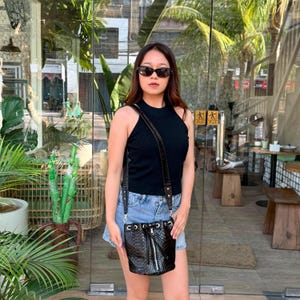 May include: A black sleeveless ribbed knit top, light blue denim shorts, and a black snakeskin-textured bucket bag with a drawstring closure and a long shoulder strap. The bag features a vertical silver-toned zipper detail on the front.
