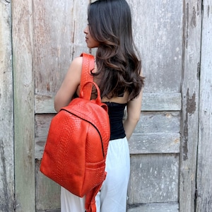 May include: A bright orange backpack featuring a textured snakeskin pattern. The bag includes a top handle, adjustable shoulder straps, and a black zipper closure that runs along the top and sides of the main compartment for secure storage.