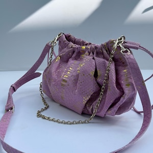 Può includere: Una borsa a sacchetto color lavanda con un motivo a pelle di serpente testurizzato e accenti in oro metallizzato. La borsa presenta una chiusura arricciata, una catena metallica argentata rimovibile e una tracolla regolabile con fibbia.