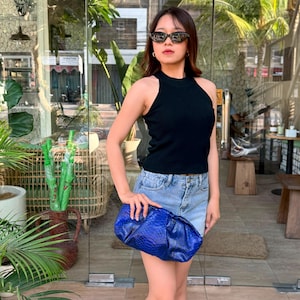 Puede incluir: Un bolso de mano azul brillante con un patrón texturizado de piel de serpiente, combinado con una camiseta negra sin mangas de canalé y pantalones cortos de mezclilla azul claro. El accesorio tiene una forma redondeada y fruncida.