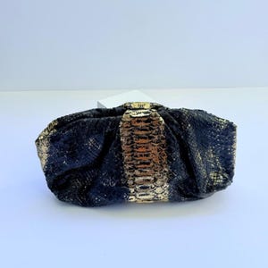 Puede incluir: Un clutch negro y dorado con un estampado de piel de serpiente. El clutch tiene forma rectangular con un diseño fruncido y un cierre dorado. Los detalles dorados añaden un toque de elegancia al accesorio.