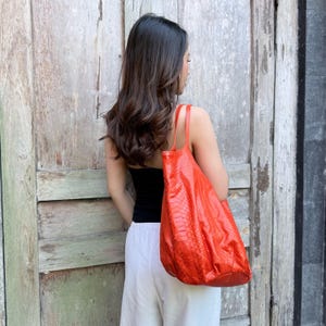 Puede incluir: Un bolso tote de color naranja brillante con un patrón texturizado de piel de serpiente. El bolso cuenta con dos correas finas para el hombro y una silueta espaciosa y holgada. El vibrante color naranja resalta la textura detallada de escamas.