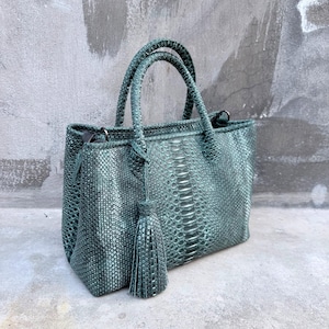 Peut inclure: Un sac à main structuré d'un ton vert sarcelle profond avec un motif texturé en peau de serpent. Le sac comprend deux poignées supérieures, une fermeture éclair sur le dessus et un pompon assorti décoratif suspendu à une poignée.