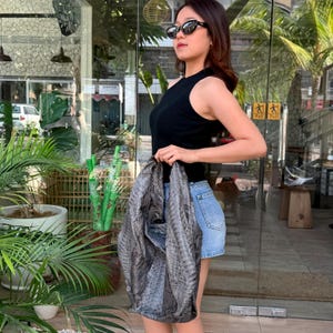 Könnte beinhalten: Ein schwarzes ärmelloses Ripp-Top, blaue Jeansshorts und eine große graue Hobo-Tasche mit Schlangenledermuster. Die Tasche hat eine strukturierte, schuppige Oberfläche mit einer lässigen Silhouette und einem breiten Riemen, seitlich getragen.