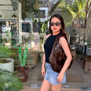 Könnte beinhalten: Eine dunkelbraune Hobo-Tasche mit Schlangenleder-Textur und breitem Schulterriemen, kombiniert mit einem schwarzen ärmellosen Ripp-Top, einem hellblauen Jeans-Minirock und einer dunkel gerahmten Cat-Eye-Sonnenbrille. Die Tasche hat eine sichelförmige Form.